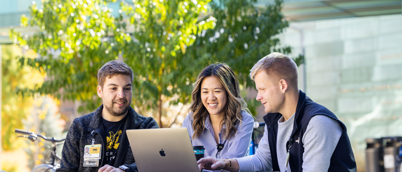 Three students sit around a laptop at an outdoor table